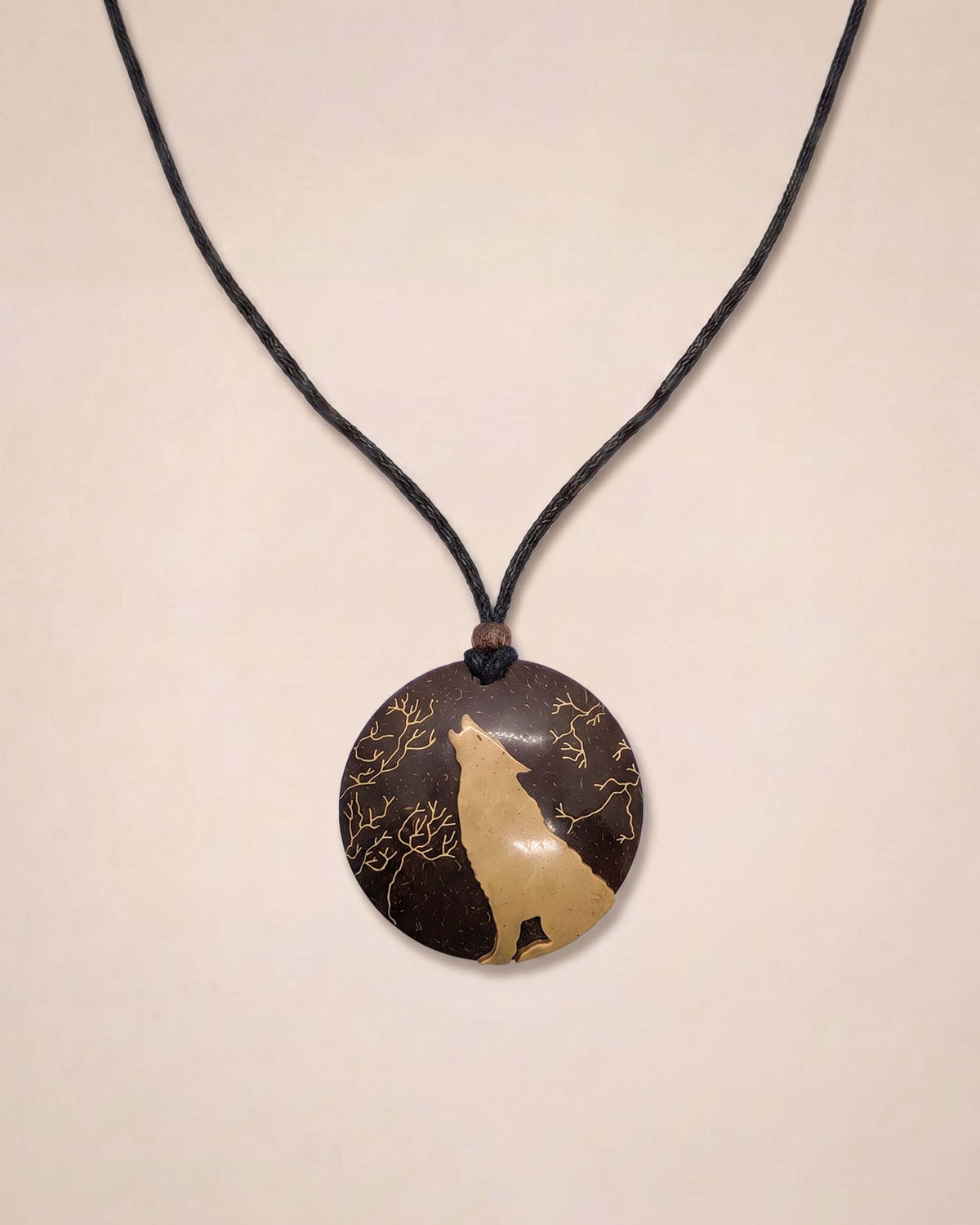 A circular pendant necklace featuring a dark brown and beige wolf silhouette design with gold branch details, suspended on a black cord against a light beige background.